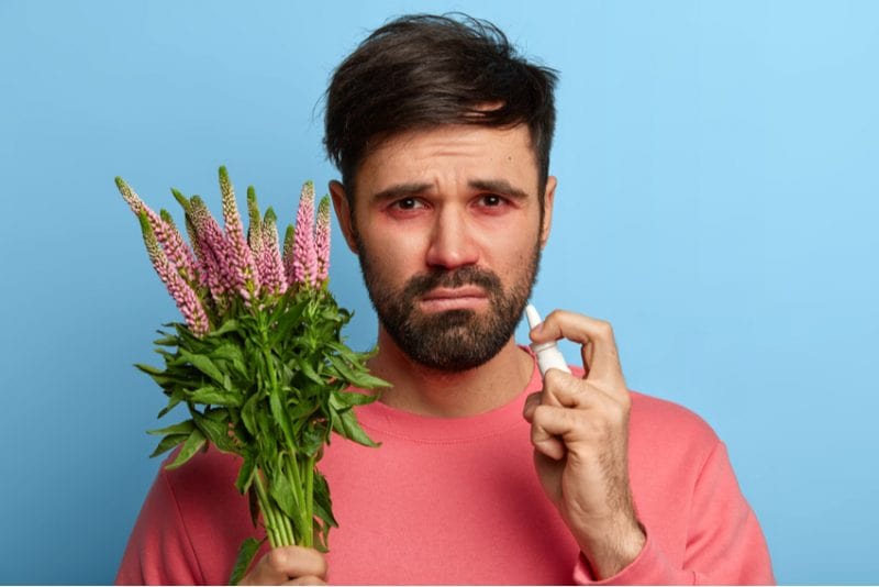 A sad looking man holding a vibrant bouquet of flowers and eye drop in his hand.
