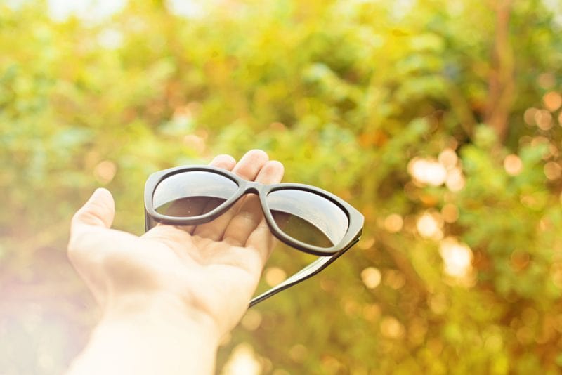 A person holding sunglasses in front of a bush.