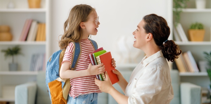 A young girl wearing glasses, holding school supplies at home with her mum